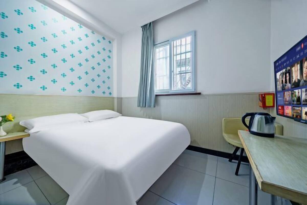 A hotel room with a bed, a small table, a chair, a window, and a wall with patterned wallpaper.