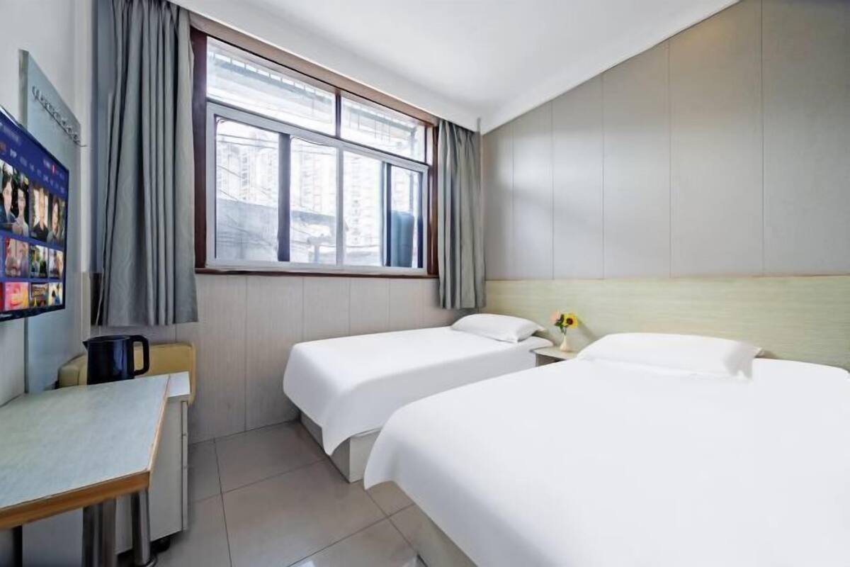 A hotel room with two beds, a desk, a window with curtains, and a wall-mounted TV.