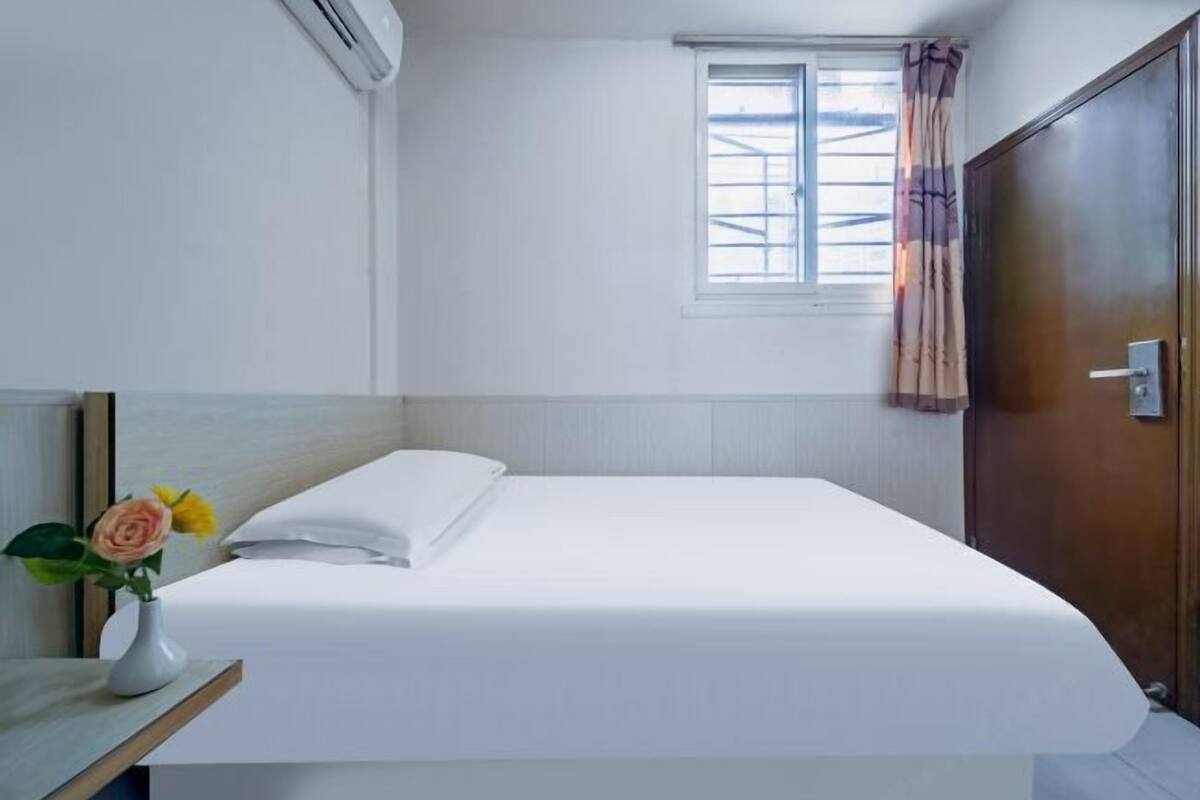 A single bed with white bedding, a small bedside table with a vase of flowers, a window with curtains, and a wooden wardrobe.