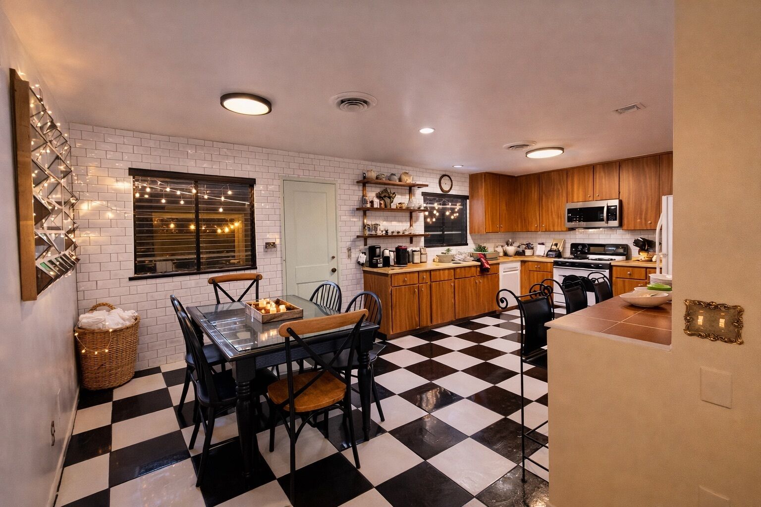 A compact kitchen with a black and white checkered floor, wooden cabinets, a microwave, and a dining area with a glass table and chairs.