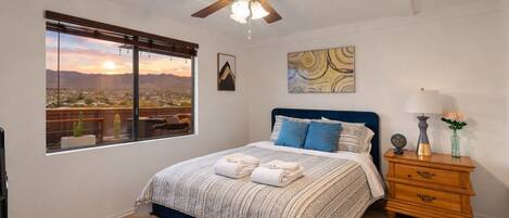 Un dormitorio con un ventanal grande, ventilador de techo, cama con ropa de cama azul, mesita de noche con lámpara y un cuadro en la pared.