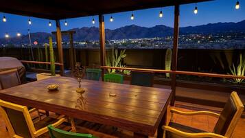 An outdoor patio with a wooden table and chairs, surrounded by cacti and desert plants, overlooking a cityscape and mountains at dusk.