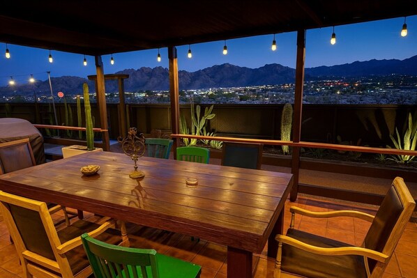 Een terras buiten met een houten tafel en stoelen, omgeven door cactussen en woestijnplanten, met uitzicht op een stadslandschap en bergen in de schemering.