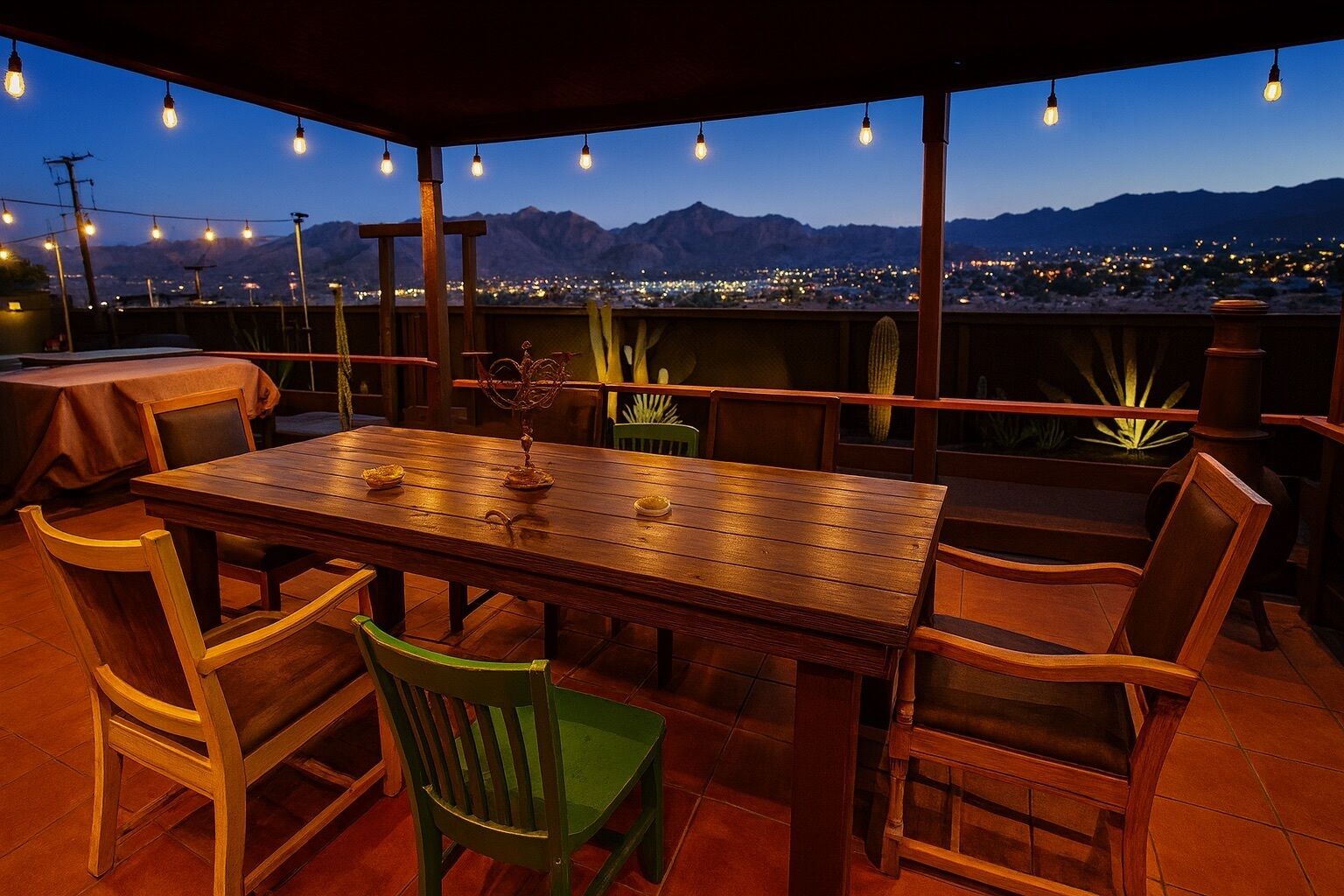 An outdoor patio with a wooden table and chairs, string lights, and a view of a cityscape and mountains at dusk.