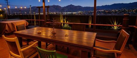 An outdoor patio with a wooden table and chairs, string lights, and a view of a cityscape and mountains at dusk.