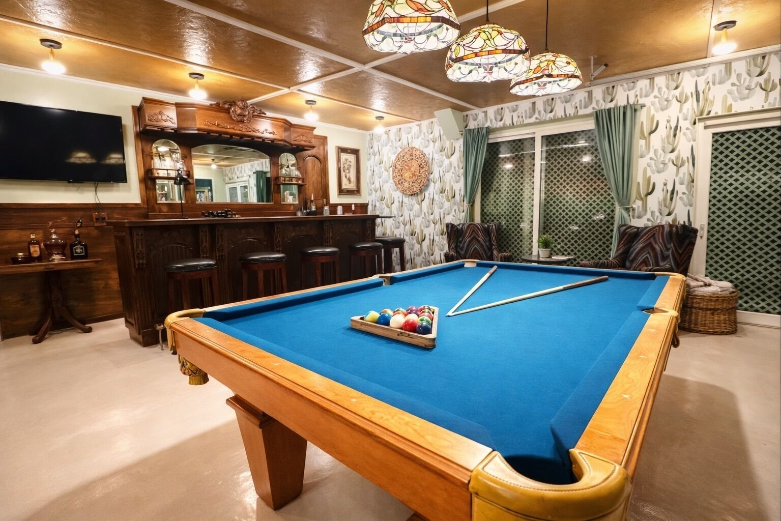 A pool table with a blue felt surface, surrounded by wooden furniture and a bar area with a mirror and stools.