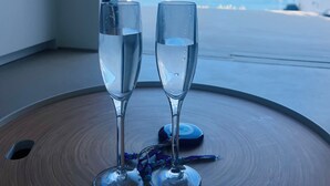 Two champagne flutes on a round wooden table with a view of a marina and boats. - ZELOS Guesthouse Milos (POLLONIAN)