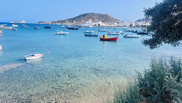 A coastal marina with boats anchored in clear blue water, surrounded by buildings and a hill in the background. - ZELOS Guesthouse Milos (POLLONIAN)