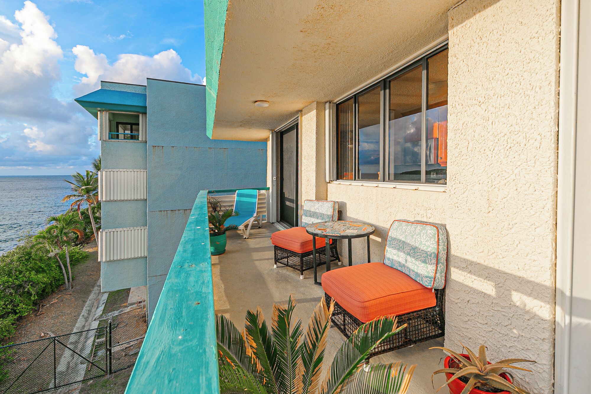A balcony with outdoor seating, a view of the ocean, and a building with a blue and beige color scheme.