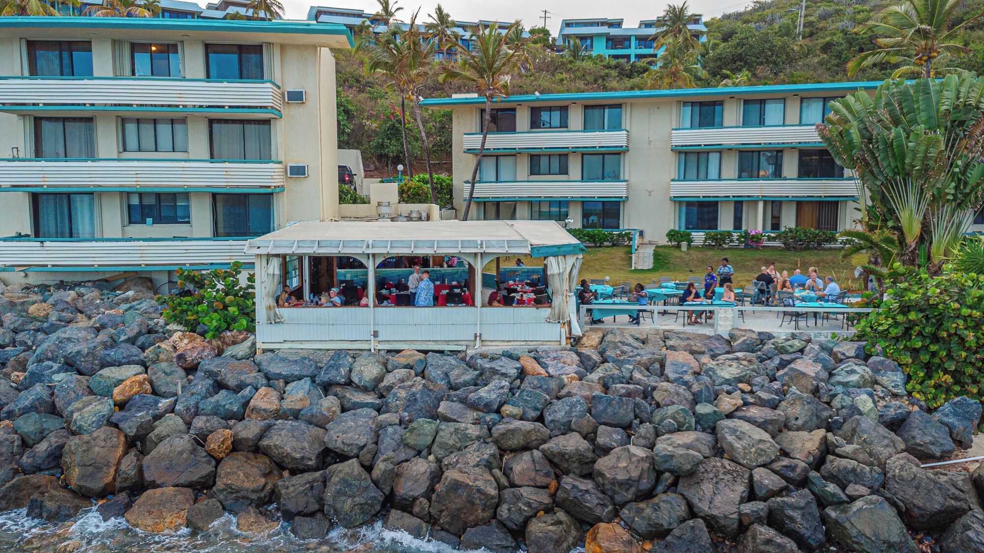 A beachfront hotel with multiple buildings, a rocky shoreline, and a dining area with tables and chairs.