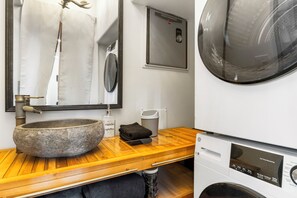 Un baño con lavamanos de piedra, espejo, dispensador de jabón, toalla y lavadora.