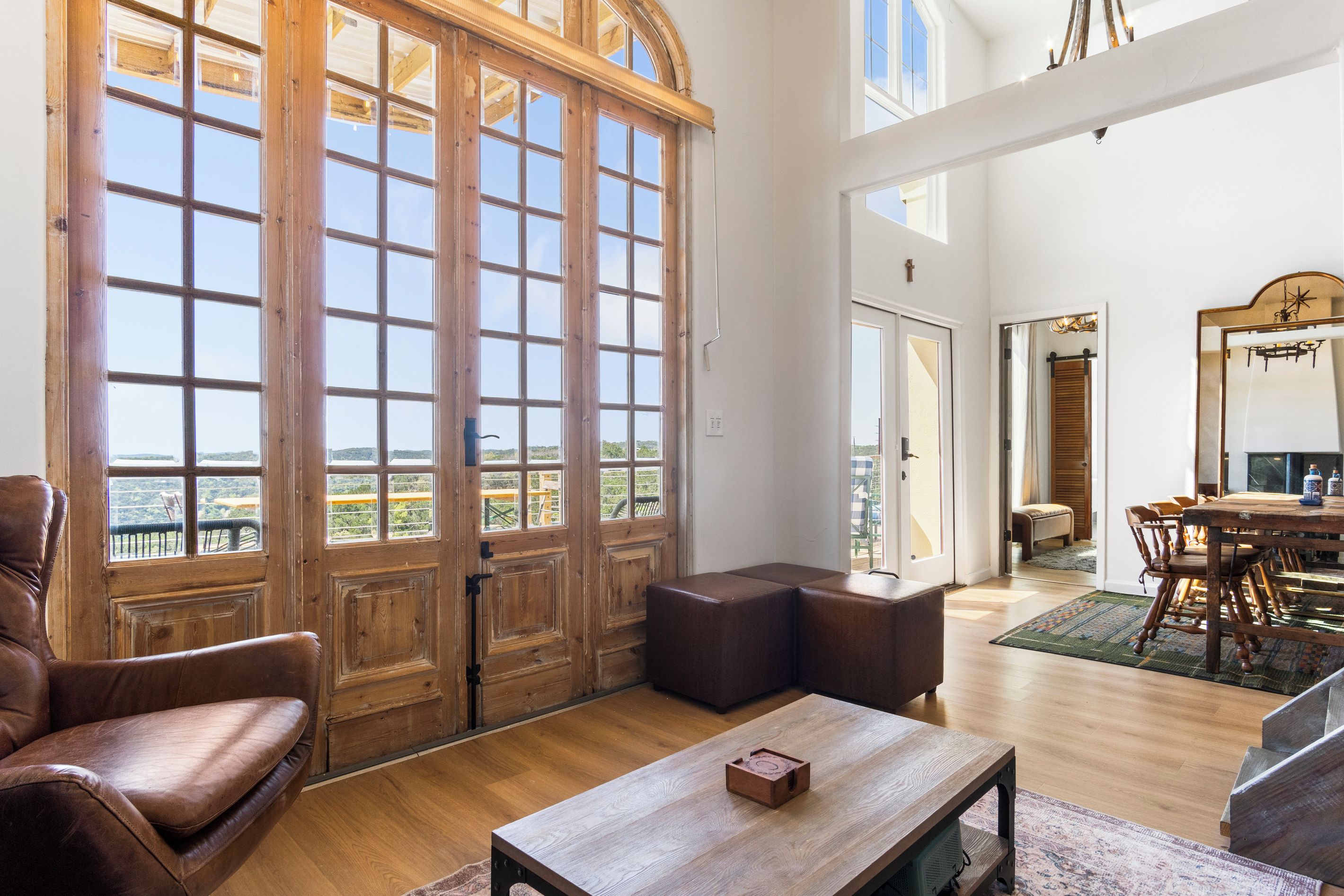Una habitación espaciosa con un gran puerta de madera que da a un balcón, un sillón de cuero, una mesa de centro de madera y un comedor con candelabro.