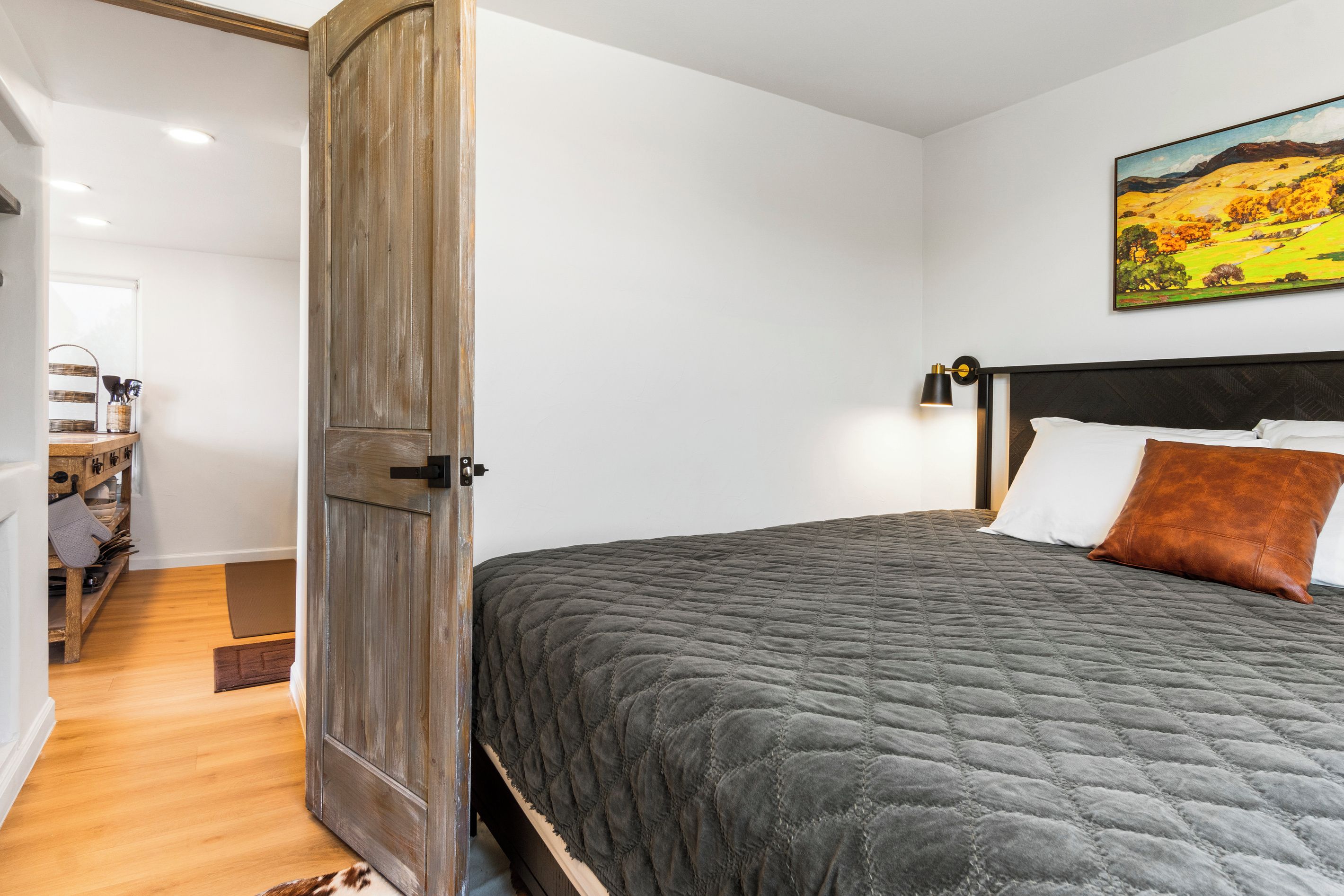 A bedroom with a large bed, a wooden wardrobe, a painting on the wall, and a small table with items on it.
