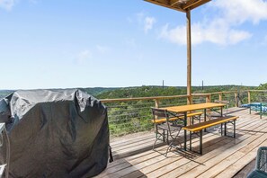 Ett trädäck med en överbyggd grillplats, picknickbord och utsikt över ett kuperat landskap.