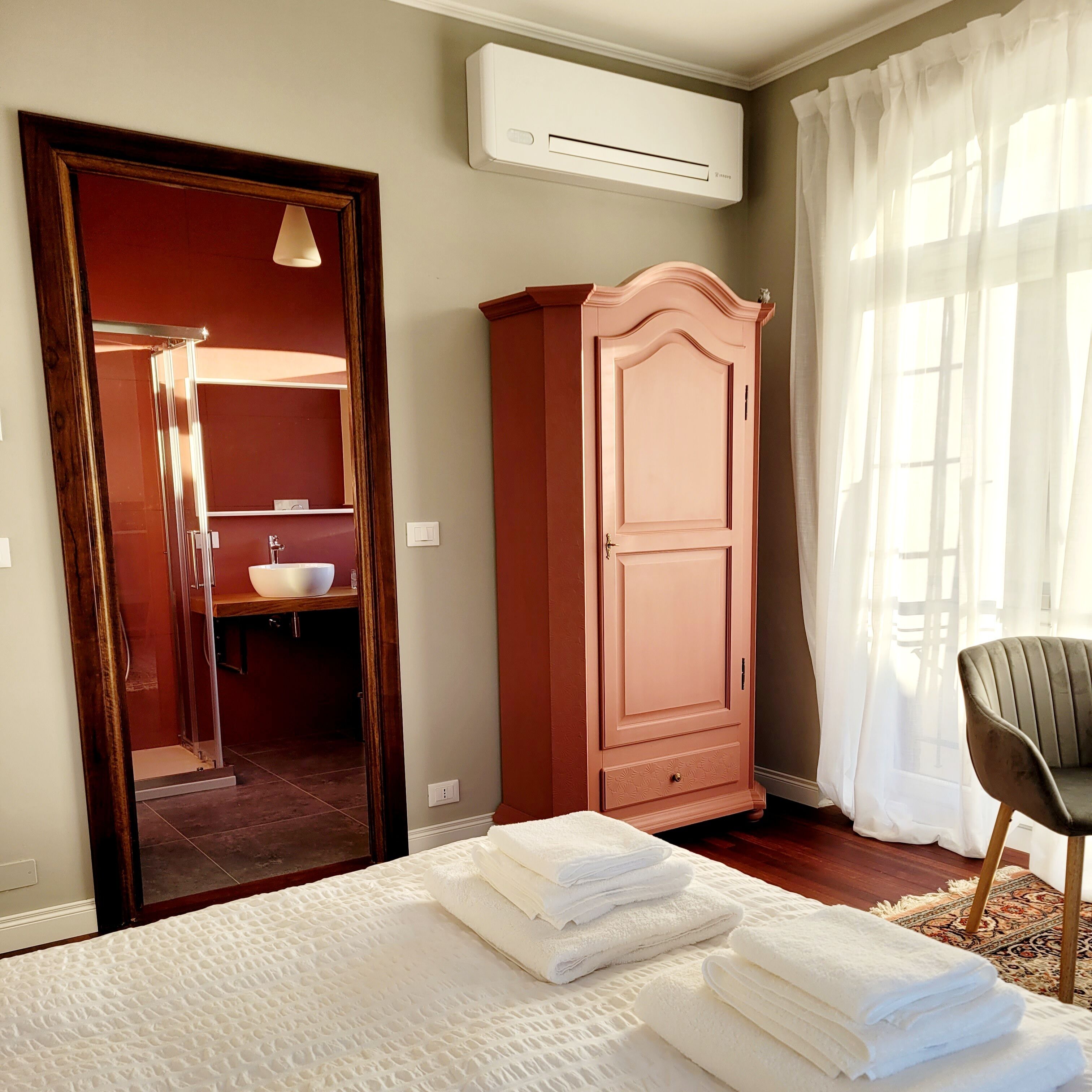 A bedroom with a bed, a wardrobe, an air conditioning unit, a bathroom with a sink, and a window with curtains.