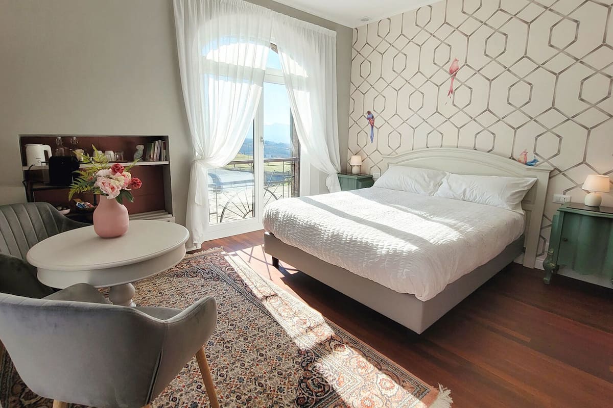 A bedroom with a large bed, a small round table with a vase of flowers, a chair, and a view of the outdoors through a window.