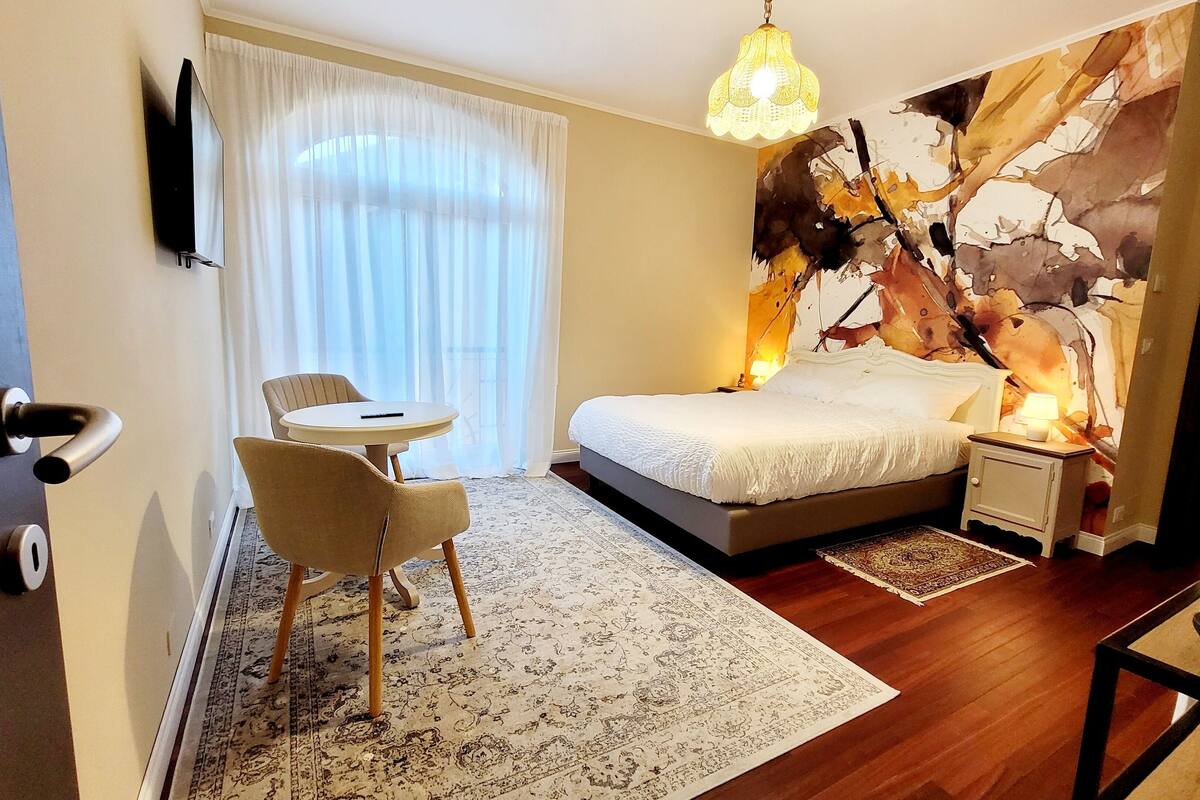 A bedroom with a bed, a chair, a small table, a rug, and a wall with a painting.