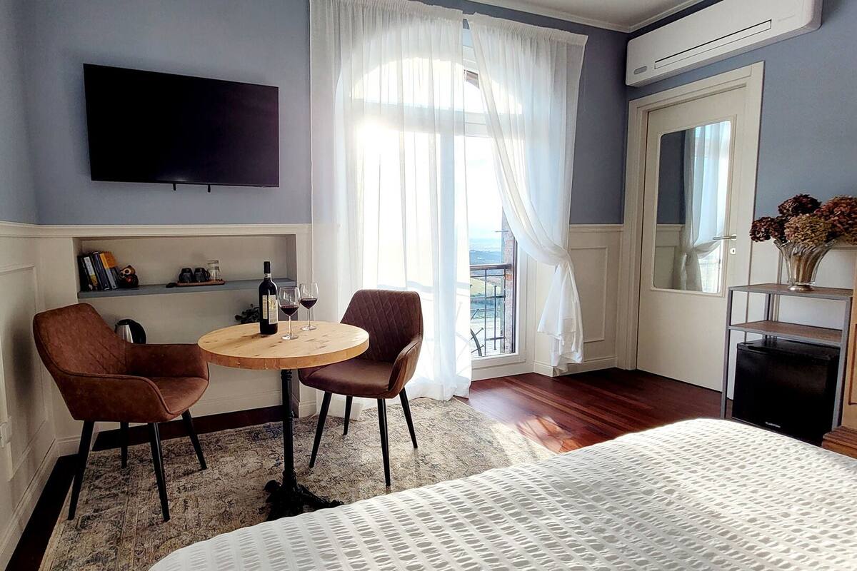 A hotel room with a bed, a TV mounted on the wall, a small round table with two chairs, a window with sheer curtains, and a view of the outside.