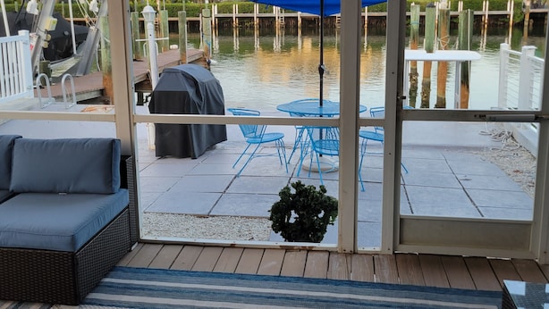 A covered outdoor seating area with a grill, blue umbrellas, and a view of the water. - Waterfront 2BR w/ Dock, Hot Tub & Easy Ocean Access – Boaters Welcome! (Marathon)