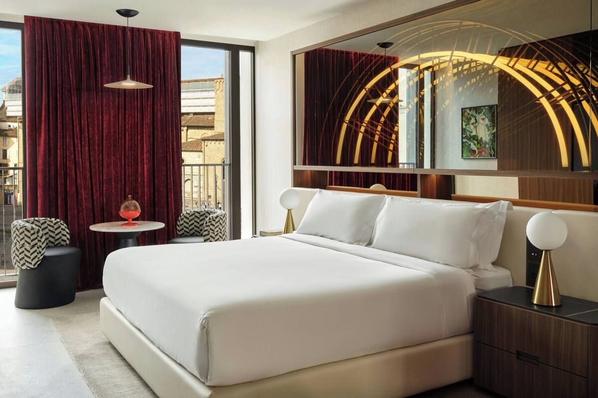 A modern hotel room with a large bed, a round table, two ottomans, a large mirror, and a view of the city.