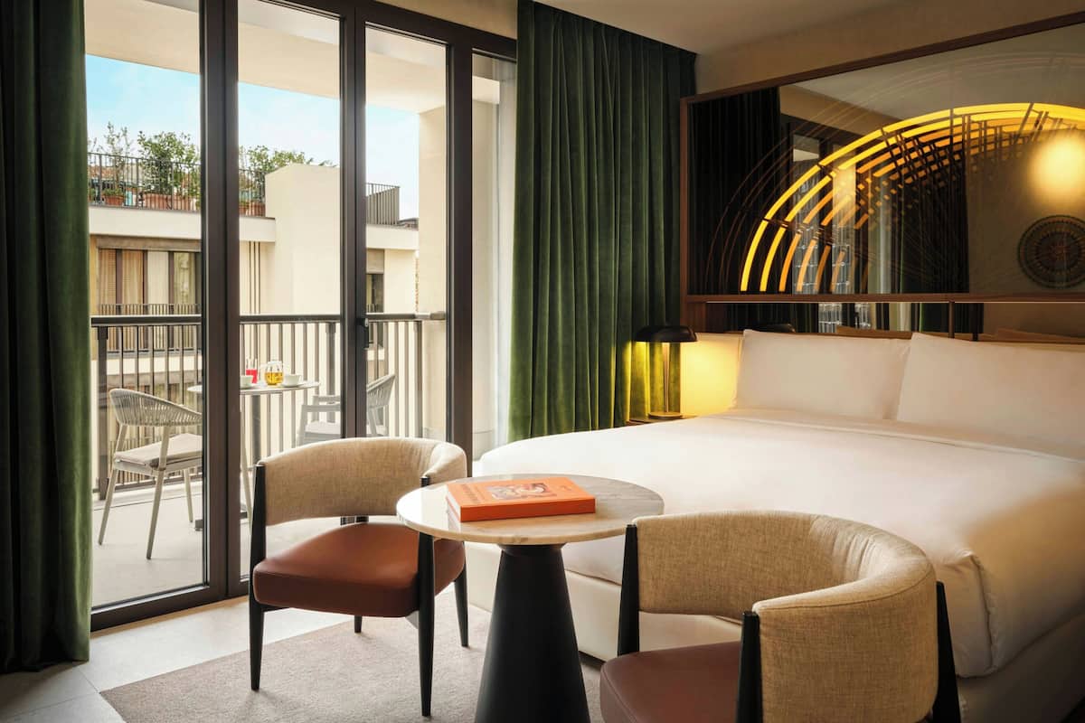 A hotel room with a bed, two chairs, a small table, a large window, and a view of the outside.