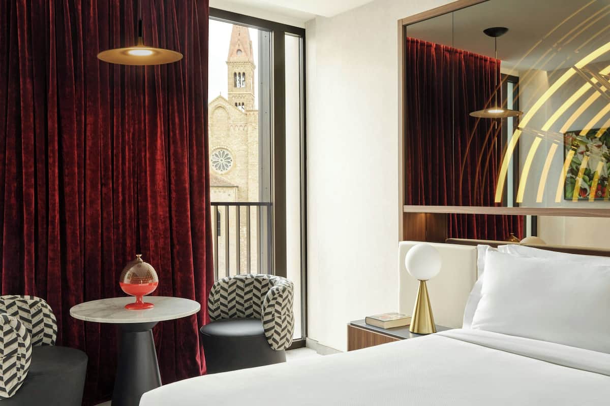 A hotel room with a bed, a round table with a red object, two chairs, a nightstand, and a large window with a view of a building.