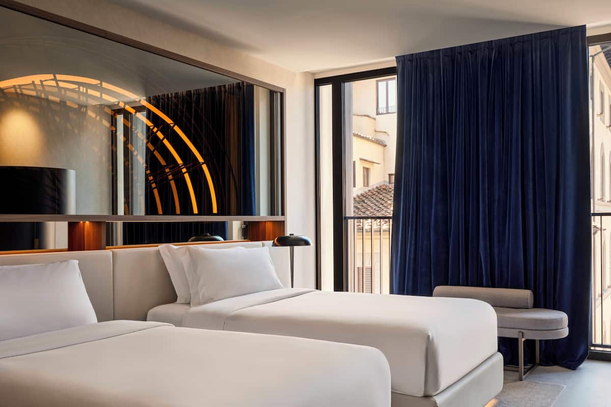 A hotel room with two beds, a large window with curtains, a small table, and a wall-mounted light fixture.
