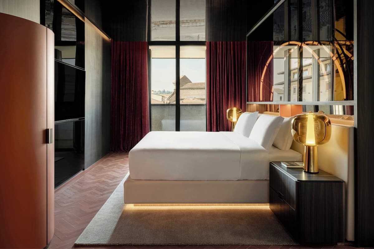 A modern hotel room with a large bed, a nightstand with a lamp, a view of buildings through large windows, and a red curtain.