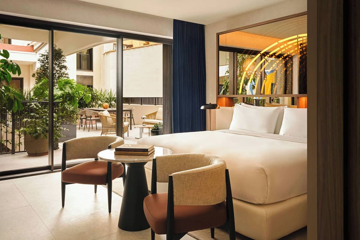 A hotel room with a large bed, two armchairs, a small table, and a view of the outdoor area through large windows.