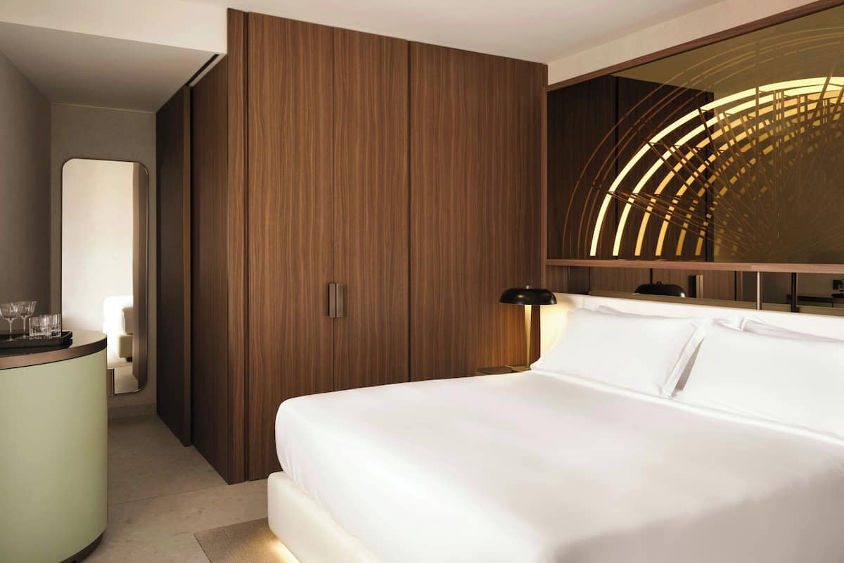 A modern hotel room with a large bed, a wooden wardrobe, a curved mirror, and a bedside table with glasses.