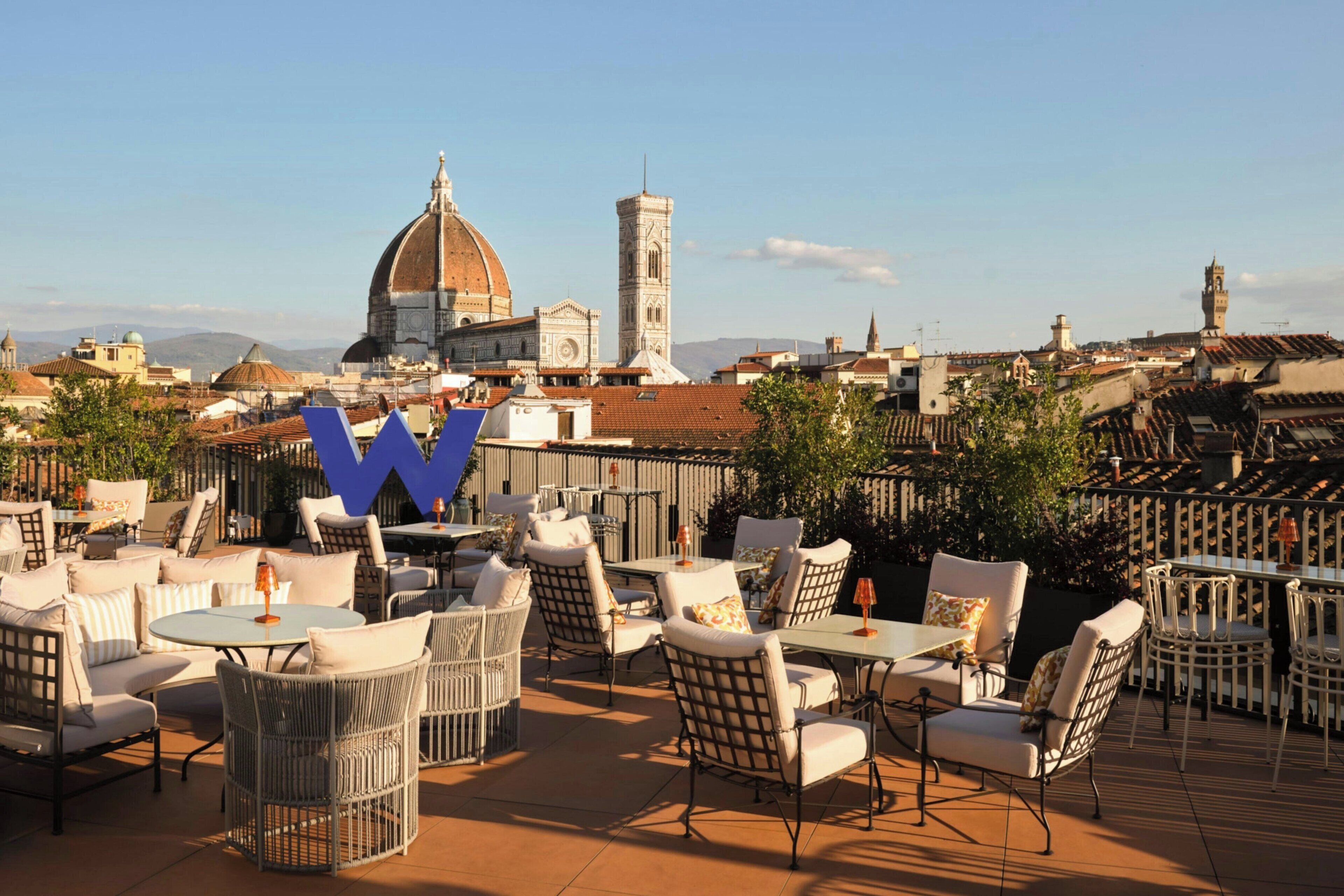 An outdoor seating area with wicker chairs and tables, overlooking a cityscape with historic buildings and a prominent dome structure.