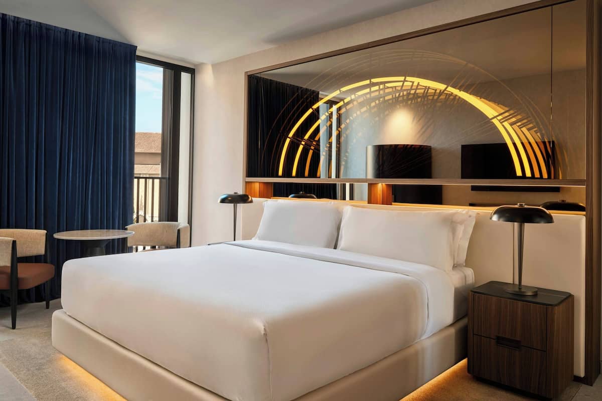 A modern hotel room with a large bed, a round mirror, a small round table, and two chairs.