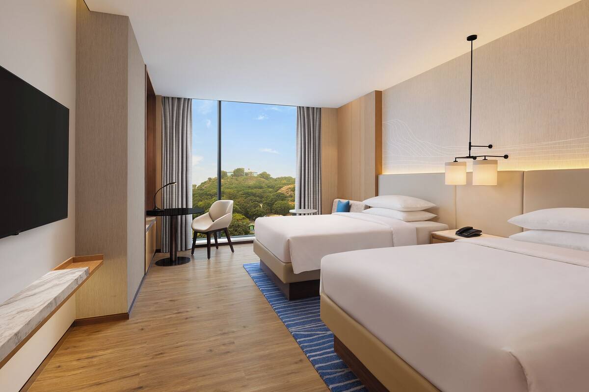 A hotel room with two beds, a desk, a chair, a TV, and a view of the outdoors.
