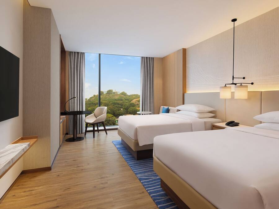 A hotel room with two beds, a desk, a chair, a TV, and a view of the outdoors.
