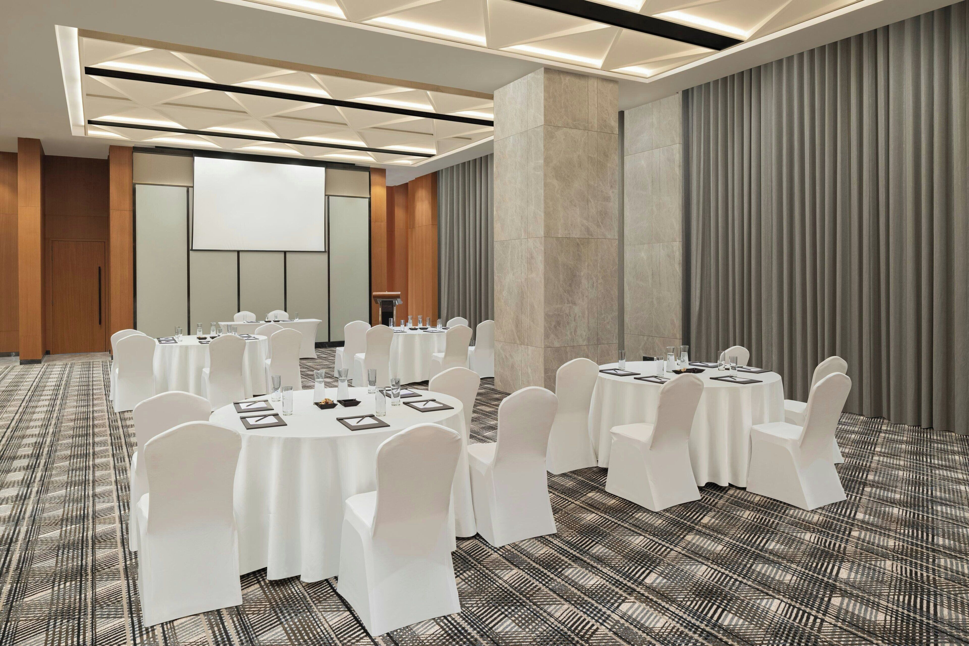 Een vergaderzaal met ronde tafels, witte stoelen en een groot scherm.