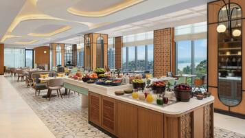 Una hall moderna di un hotel con un banco colazione centrale, zone relax e ampie finestre con vista sulla città.