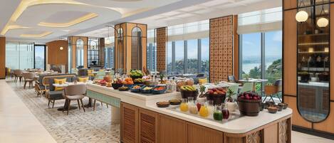 A modern hotel lobby with a central breakfast bar, seating areas, and large windows offering a city view.