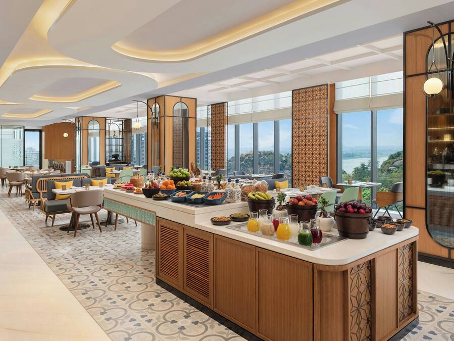 Una hall moderna di un hotel con un banco colazione centrale, zone relax e ampie finestre con vista sulla città.