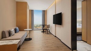 A modern hotel room with a sofa, a small table, a TV, and a view through large windows.