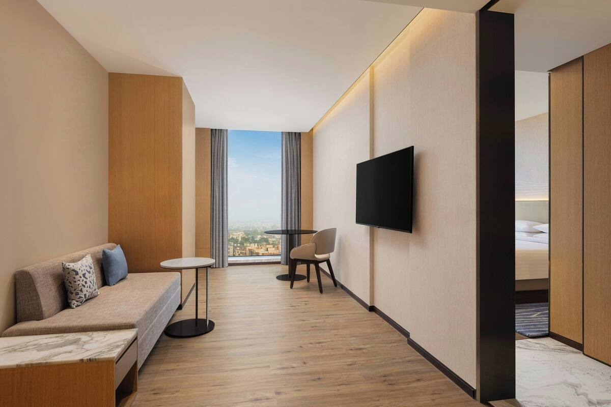 A modern hotel room with a sofa, a small table, a TV, and a view through large windows.