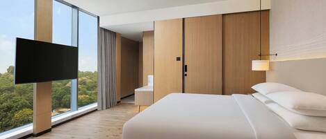 Una habitación de hotel moderna con una cama grande, un televisor de pantalla plana montado en un panel de madera, un escritorio con una silla y un ventanal con vistas al exterior.