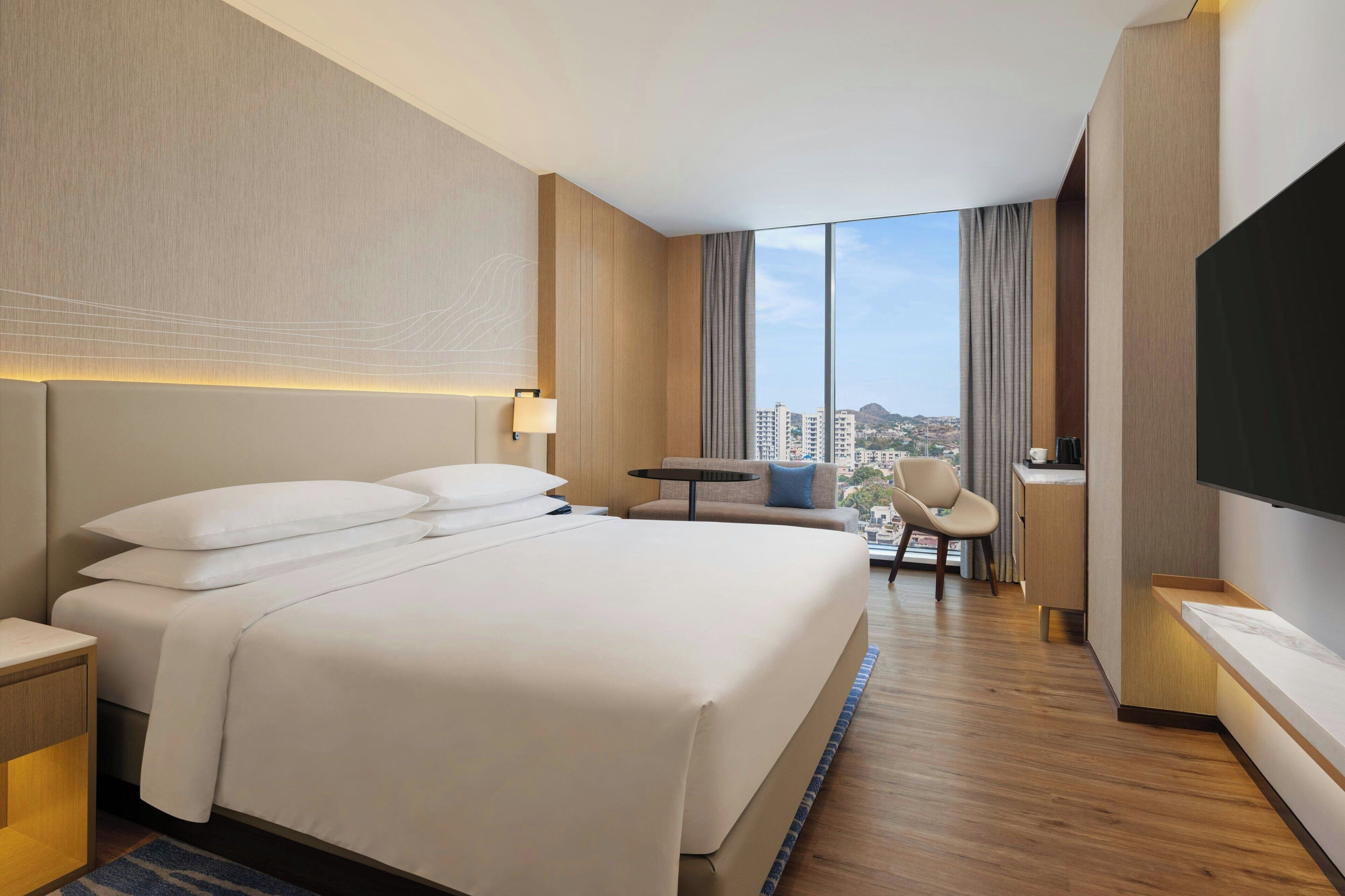 Ein Hotelzimmer mit einem großen Bett, einem Schreibtisch, einem Sessel, einem Fernseher und Blick auf die Stadt.