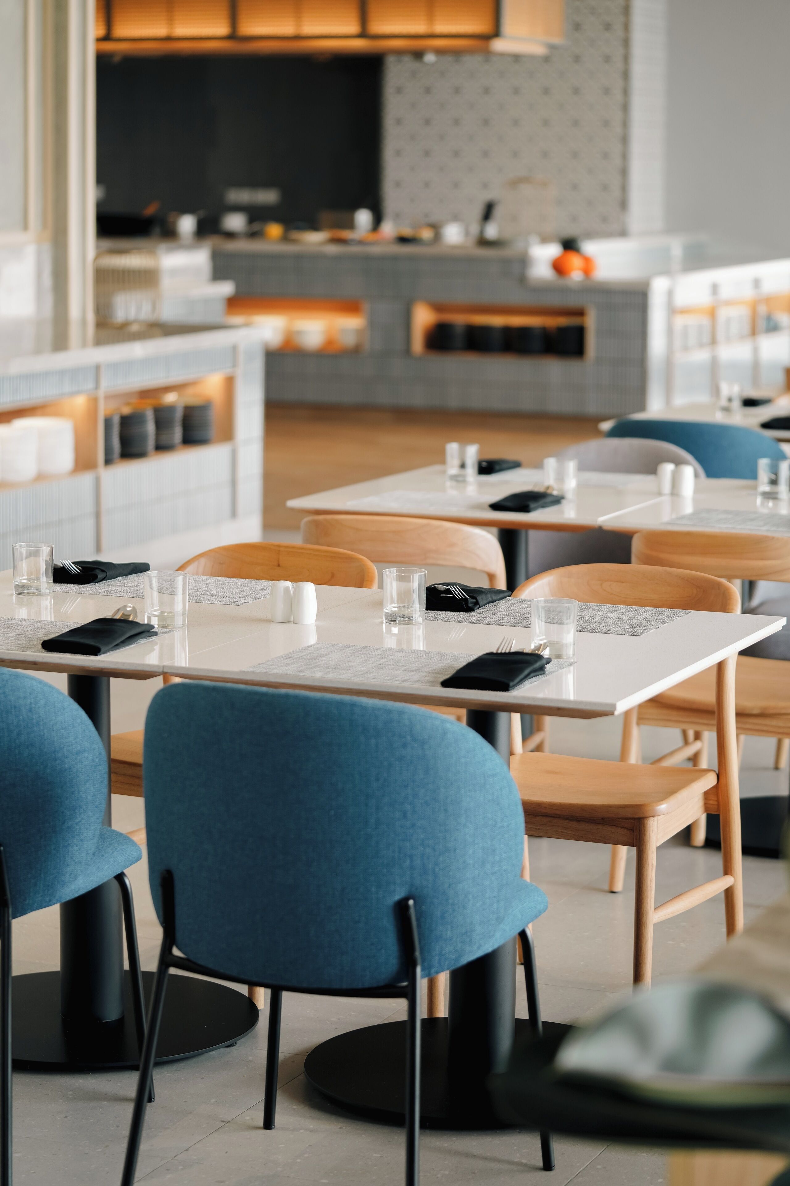 Un restaurante moderno con mesas y sillas ordenadas, un diseño interior limpio y minimalista, y un área de cocina visible al fondo.