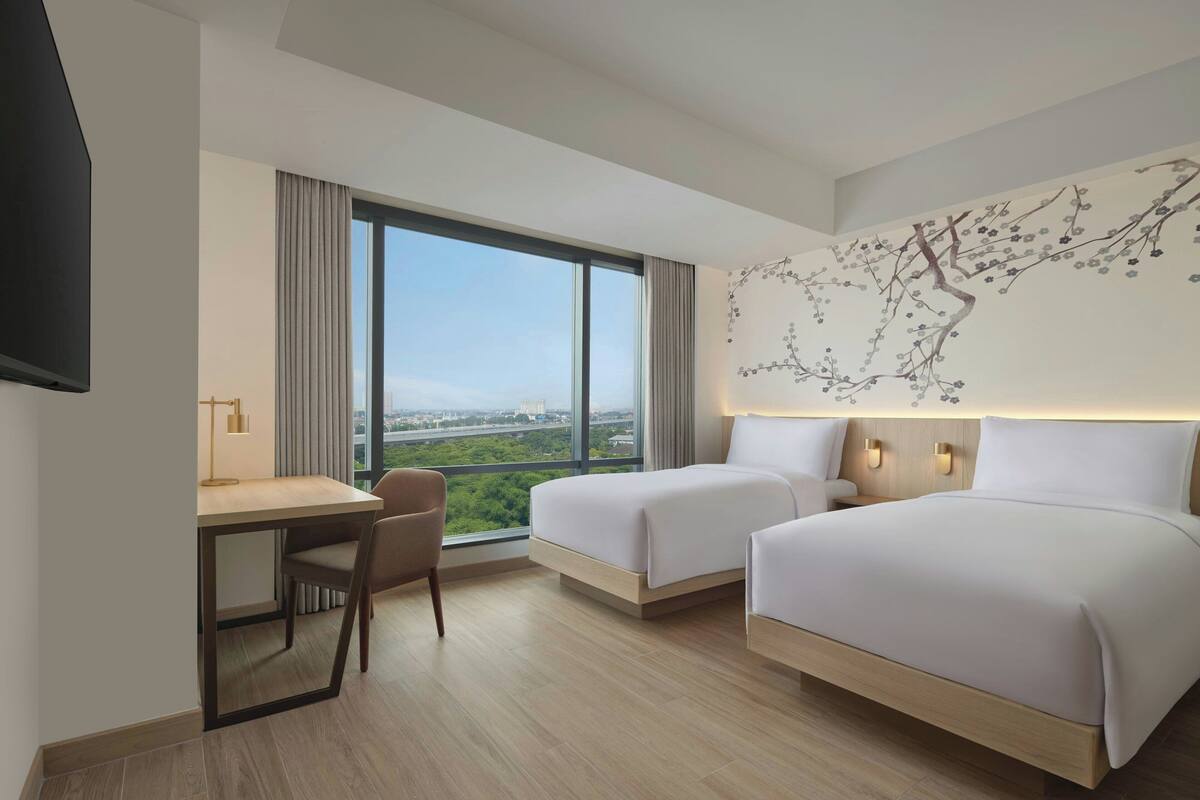 Une chambre d’hôtel avec deux lits, un bureau et une chaise. Une grande fenêtre offre une vue sur la ville.