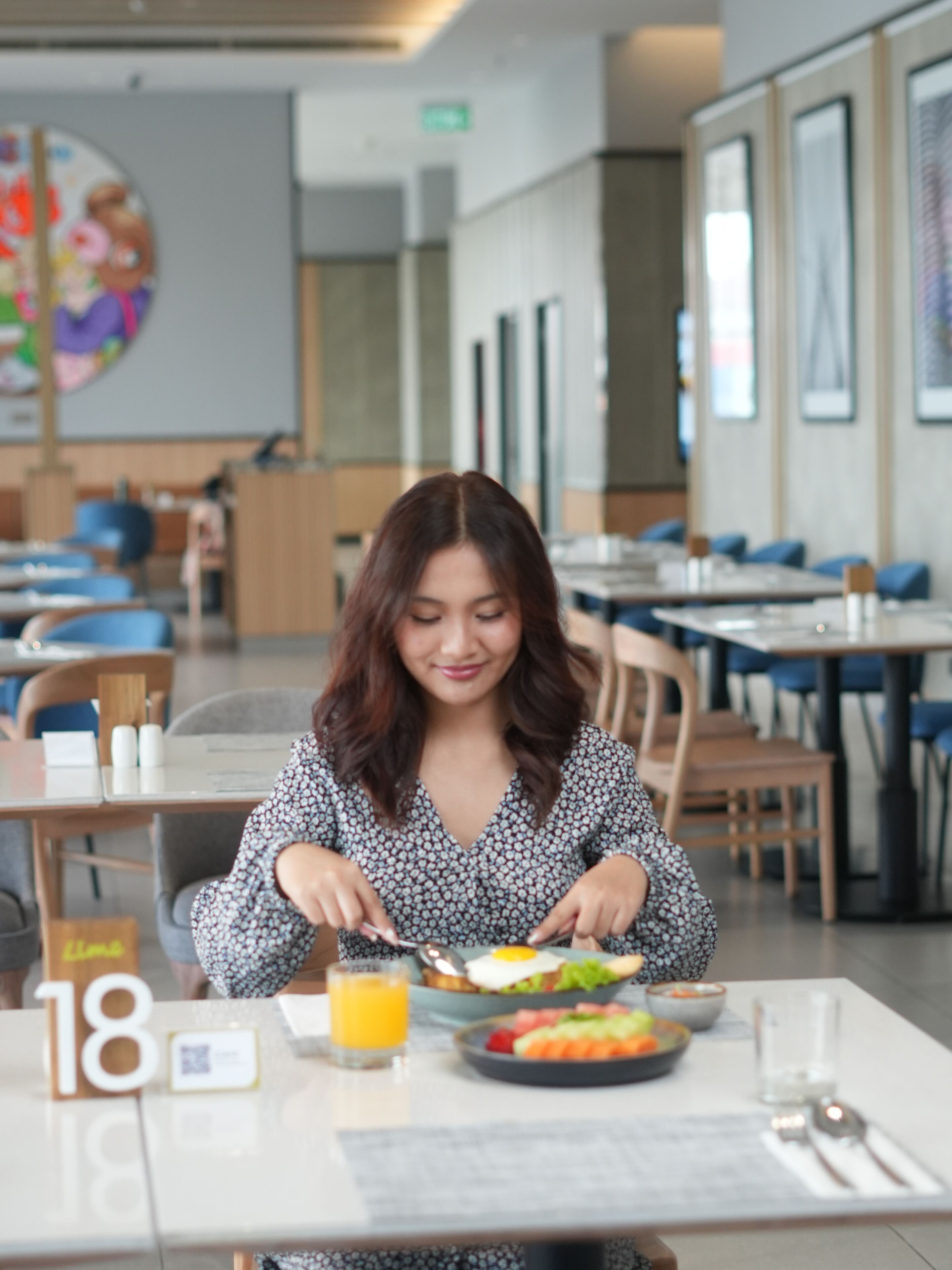 A person seated at a table with a meal, in a modern dining area with multiple tables and chairs, and a large wall art piece.