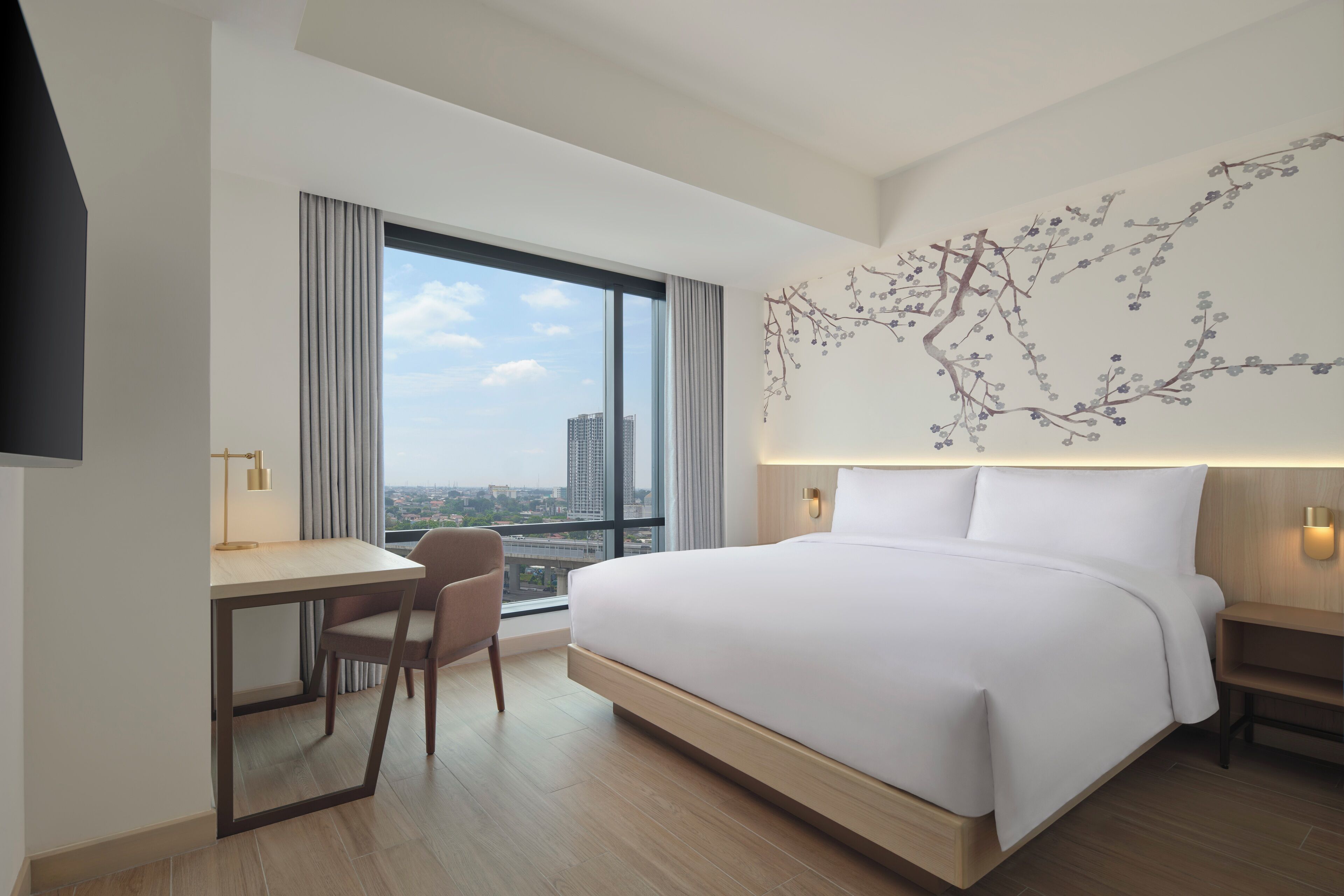 A modern hotel room with a large bed, a desk, and a chair. There is a large window with a view of the city.