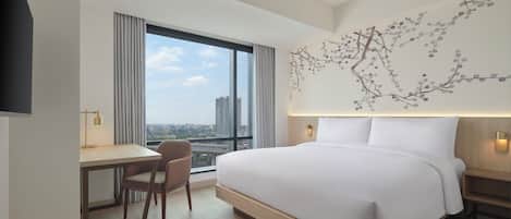 A modern hotel room with a large bed, a desk, and a chair. There is a large window with a view of the city.