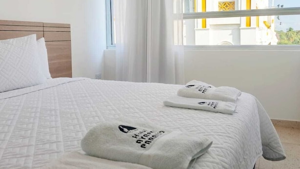 A hotel room with a large bed, white bedding, and two towels with text. A window offers a view of a building with yellow and white architecture. - Hotel Ayapel Paraiso (Ayapel)
