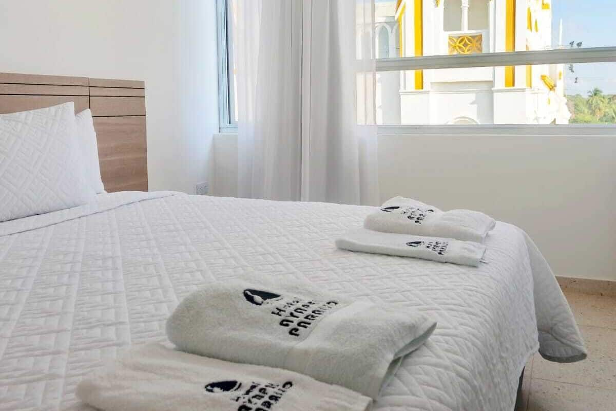 Ett hotellrum med en stor säng, vita sängkläder och två handdukar med tryck. Ett fönster ger utsikt över en byggnad med gul och vit arkitektur.