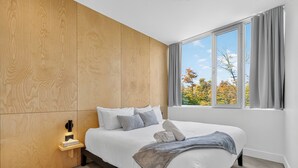A modern bedroom with a large bed, a nightstand, a wall-mounted light, and a window with curtains.